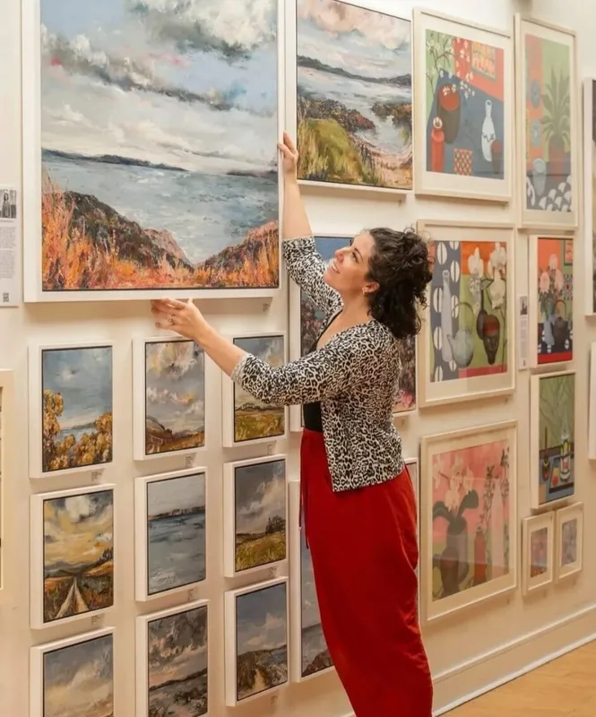 Virginia reaching up to hang a painting on a gallery wall covered in framed work