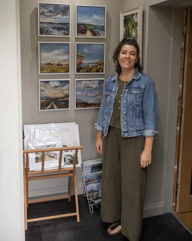 Virginia standing in a doorway in a denim jacket, small framed landscapes on the wall beside her