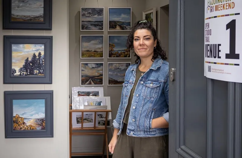 Virginia smiling in the open front door of her home, a Venue 1 sign on the door and framed paintings in the hall