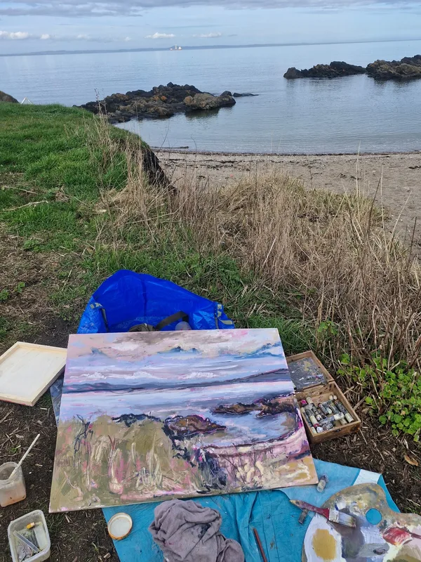 A painting of the shore propped up on a blue sheet among the rocks and grass it shows, with the sea behind