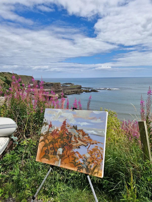 A painting on a small easel among pink willowherb on the clifftop, the sea and harbour behind it