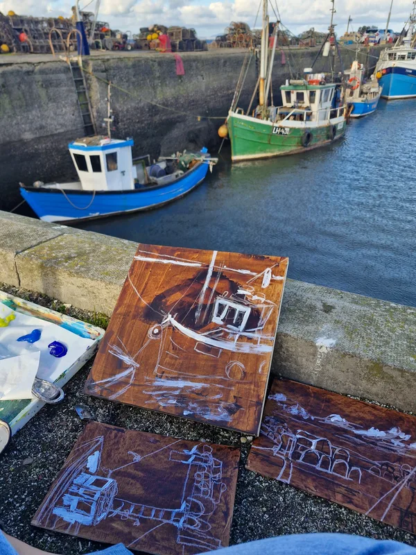 Three small paintings of fishing boats laid out on the harbour wall above the boats themselves