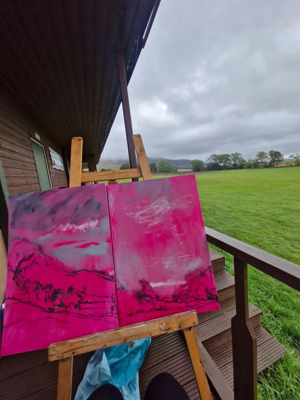 A pink painted canvas on an easel on a decking, a green field behind