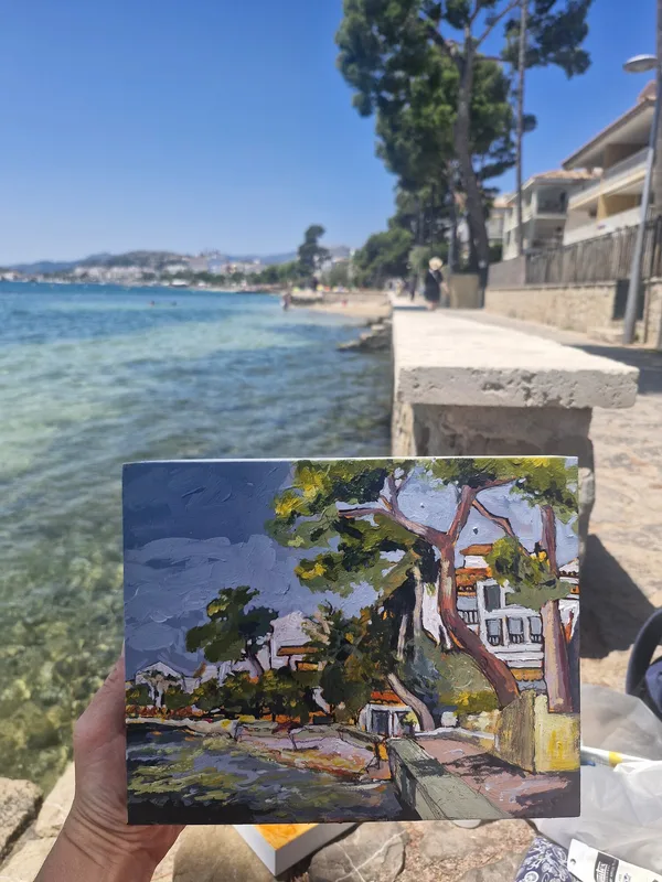 A small painting of white villas and palms held up in front of the seafront path it shows, the sea and hills beyond