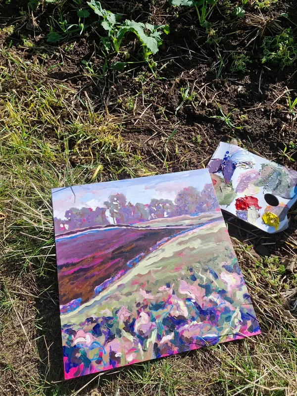 A painting of purple and green foliage on the grass beside a riverbank, with a palette of paints