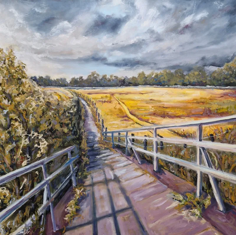 A long footbridge with white railings crossing towards golden fields and a line of trees, under a grey cloudy sky
